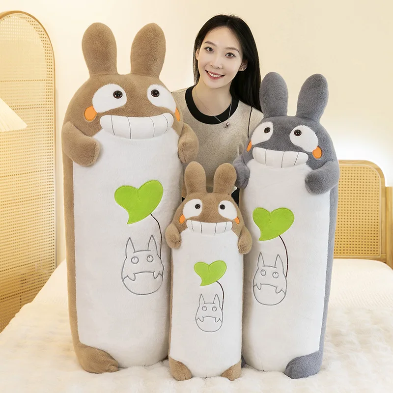 Totoro-kudde med stort leende, söt tecknad karaktärsdesign och mjuk lång form 24 Tre långörade gosedjur i tan och grått visas i upprätt form framför en person.