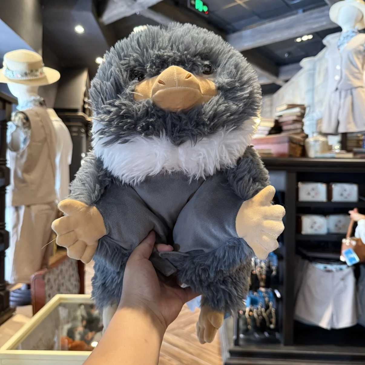 Niffler gosedjur som plyschkudde och karaktärsdocka, souvenir från Fantastic Beasts 19 Lurvig gråvit figur med gulorange näbb och händer framför butikshyllor.