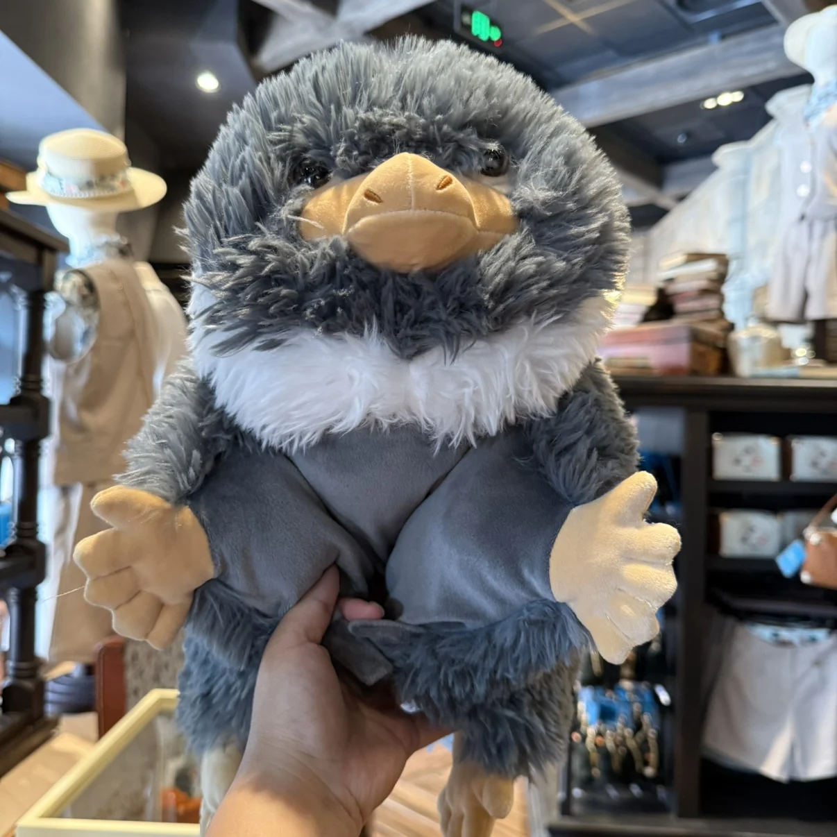 Niffler gosedjur som plyschkudde och karaktärsdocka, souvenir från Fantastic Beasts 18 Lurvig grå Nifflerfigur med ljus bröstpäls och gulorange händer och fötter.