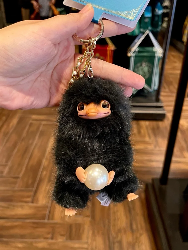 Svart Niffler-nyckelring med boll, hängande väskdekoration från Magic Academy 19 Svart, lurvig plyschfigur med orange näbb som håller en blank kula och har nyckelringskedja.